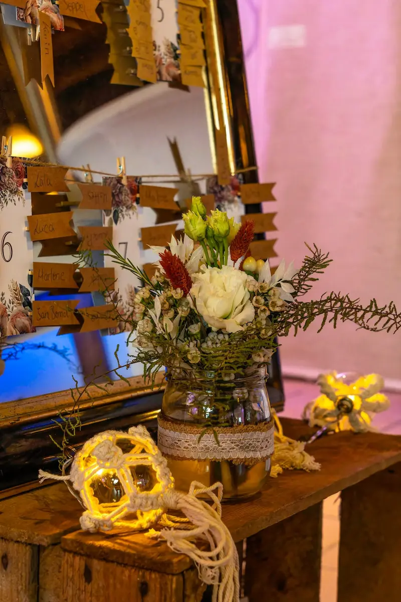 "Ein hölzerner Tisch mit einem großen Spiegel, an dem Namenskärtchen für einen Hochzeit-Sitzplan befestigt sind. Im Vordergrund stehen ein herbstlicher Blumenstrauß in einem Spitzenglas und leuchtende Makramee-Kugeln."