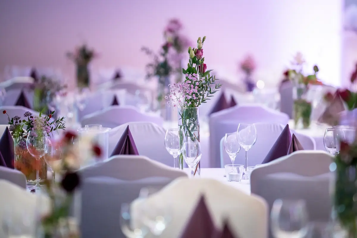 Festsaal mit festlich dekorierten Tischen. Im Fokus steht eine schlanke Glasvase mit rosa und violetten Blumen sowie grünem Beiwerk. Die Stühle sind mit weißen Hussen überzogen. Auf den Tischen stehen Weingläser und violette, spitz gefaltete Servietten. Der Hintergrund ist in sanftes, lila-rosa Licht getaucht.
